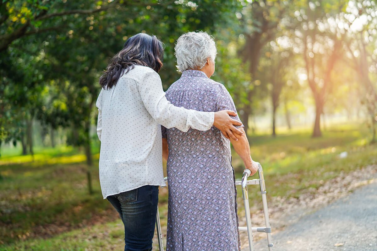 A caregiver walking outdoors beside an older woman using a walking frame