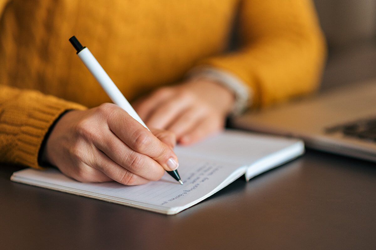 A person writing notes in a notebook beside a laptop